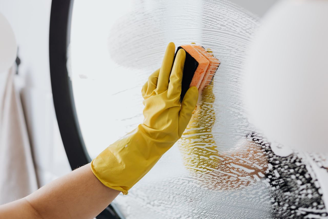 about-02 Personne anonyme portant des gants en caoutchouc jaunes et utilisant une éponge simple pour nettoyer un miroir dans une salle de bain